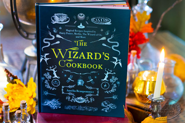 Wizarding Cookbook displayed on a table with candles, apples, glass bottles, and leaves
