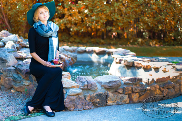 Young Woman wearing a scarf and hat sits on a stone fountain enjoying a cup of tea from a porcelain teacup on a fall day