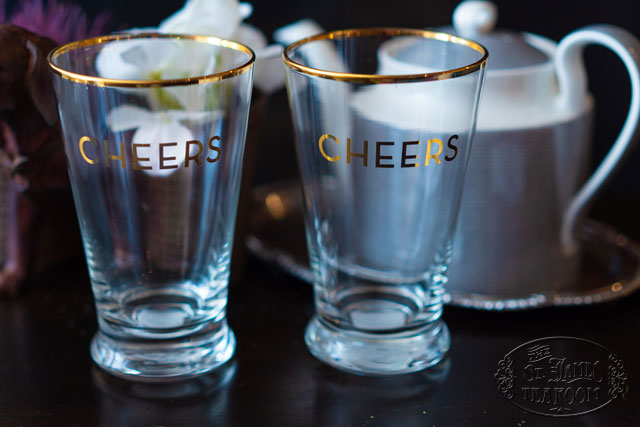 Glass pint glasses decorated with the word "Cheers" on a wooden table with a tea pot and flowers in the background